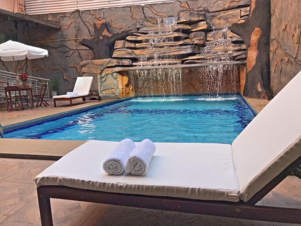 A pool with a waterfall feature, a lounge chair with towels, and a table with chairs.
