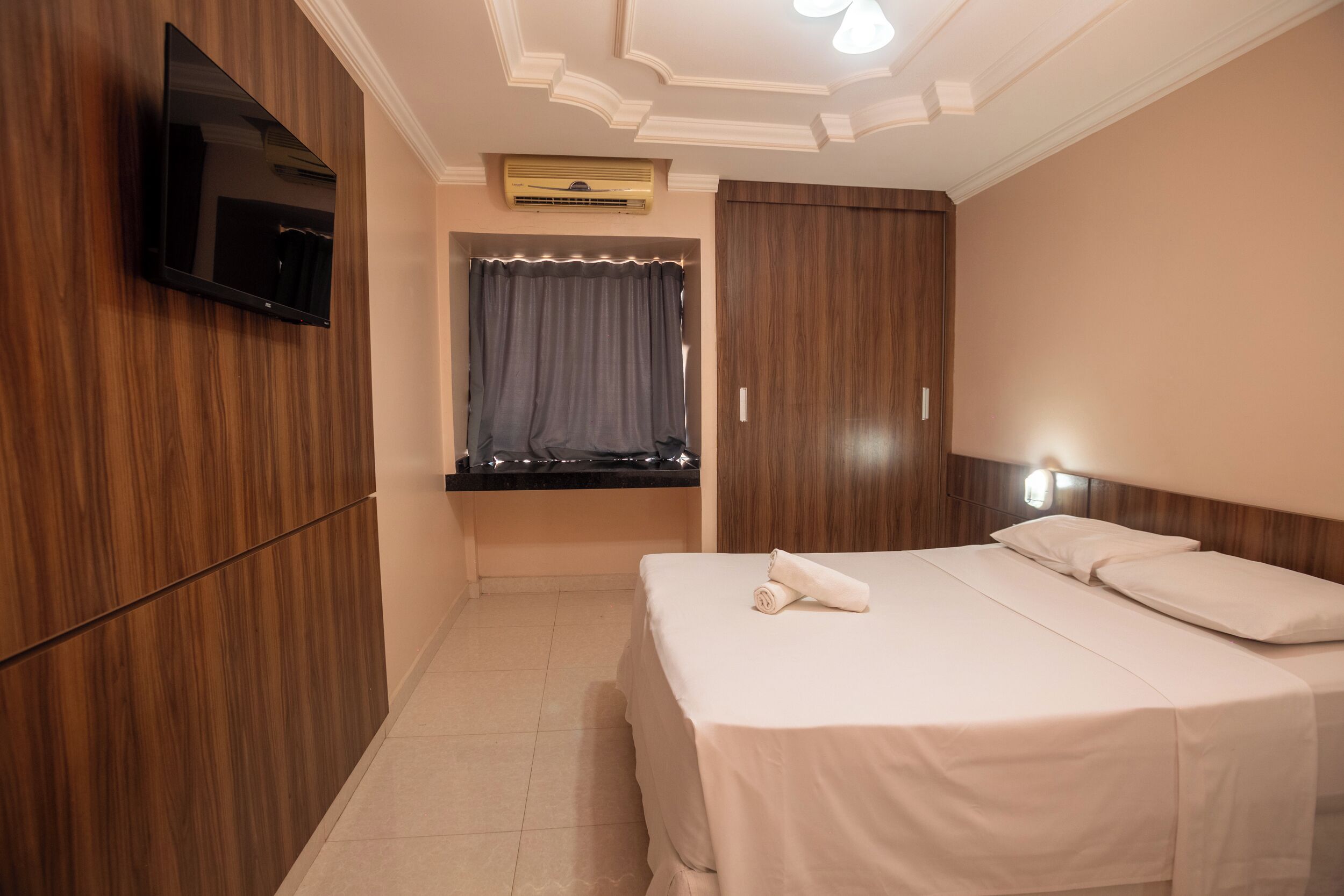 Habitación de hotel con una cama, un televisor de pantalla plana montado en la pared, una ventana con cortinas y un armario de madera.