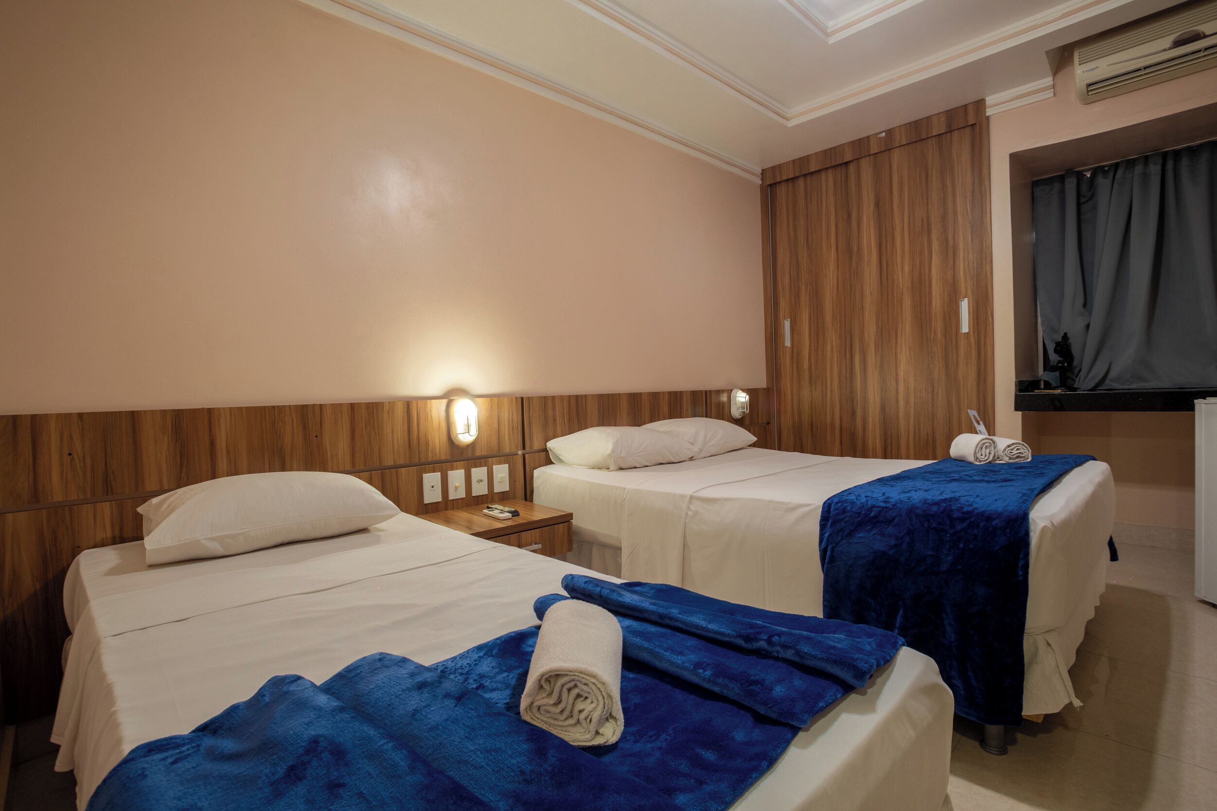 Een hotelkamer met twee bedden, elk voorzien van een blauwe deken en een opgerolde handdoek. Er is een houten hoofdbord, een nachtkastje met een lamp, en een wandgemonteerde airconditioning.