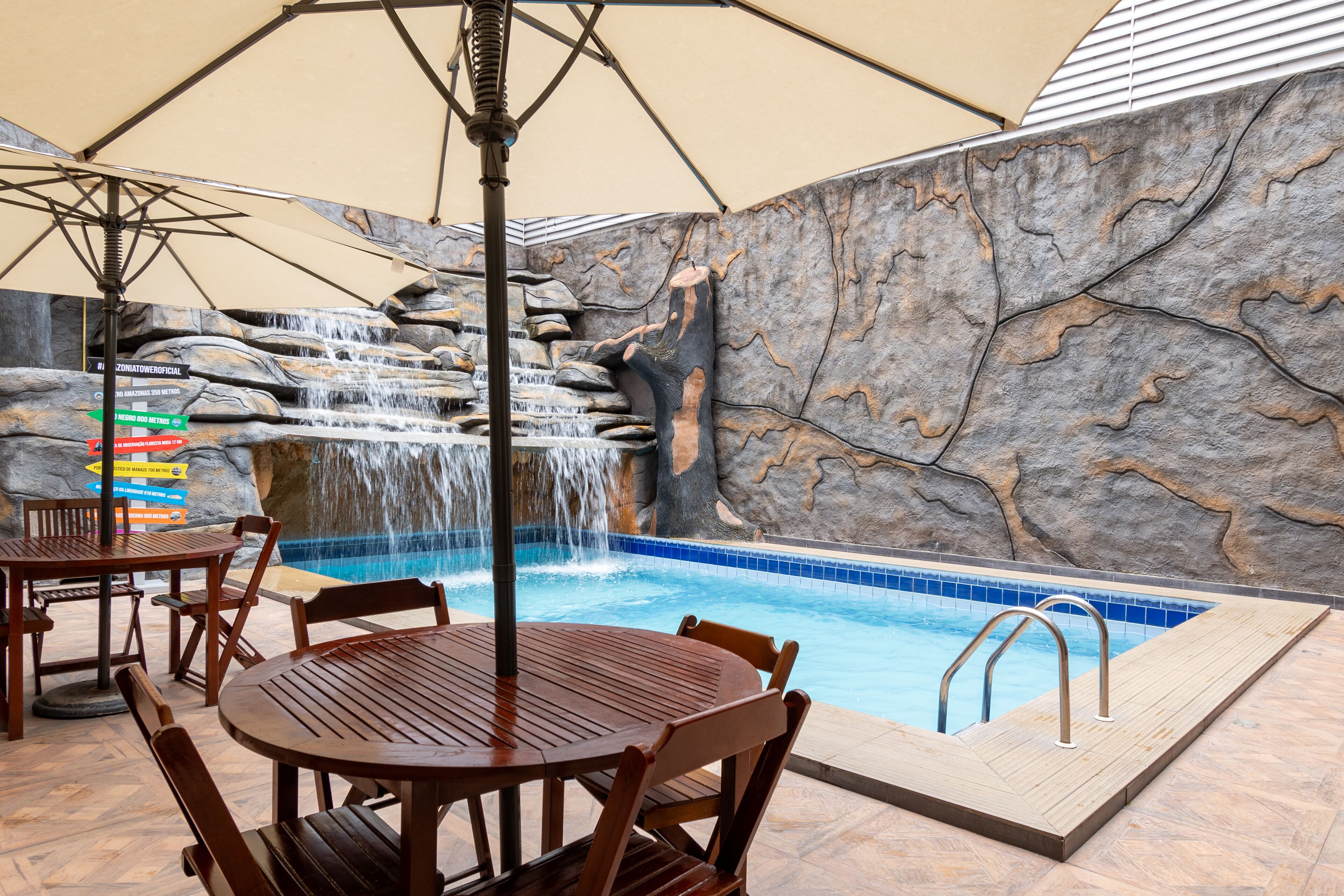 An indoor pool area with a waterfall feature, wooden poolside furniture, and a stone wall backdrop.