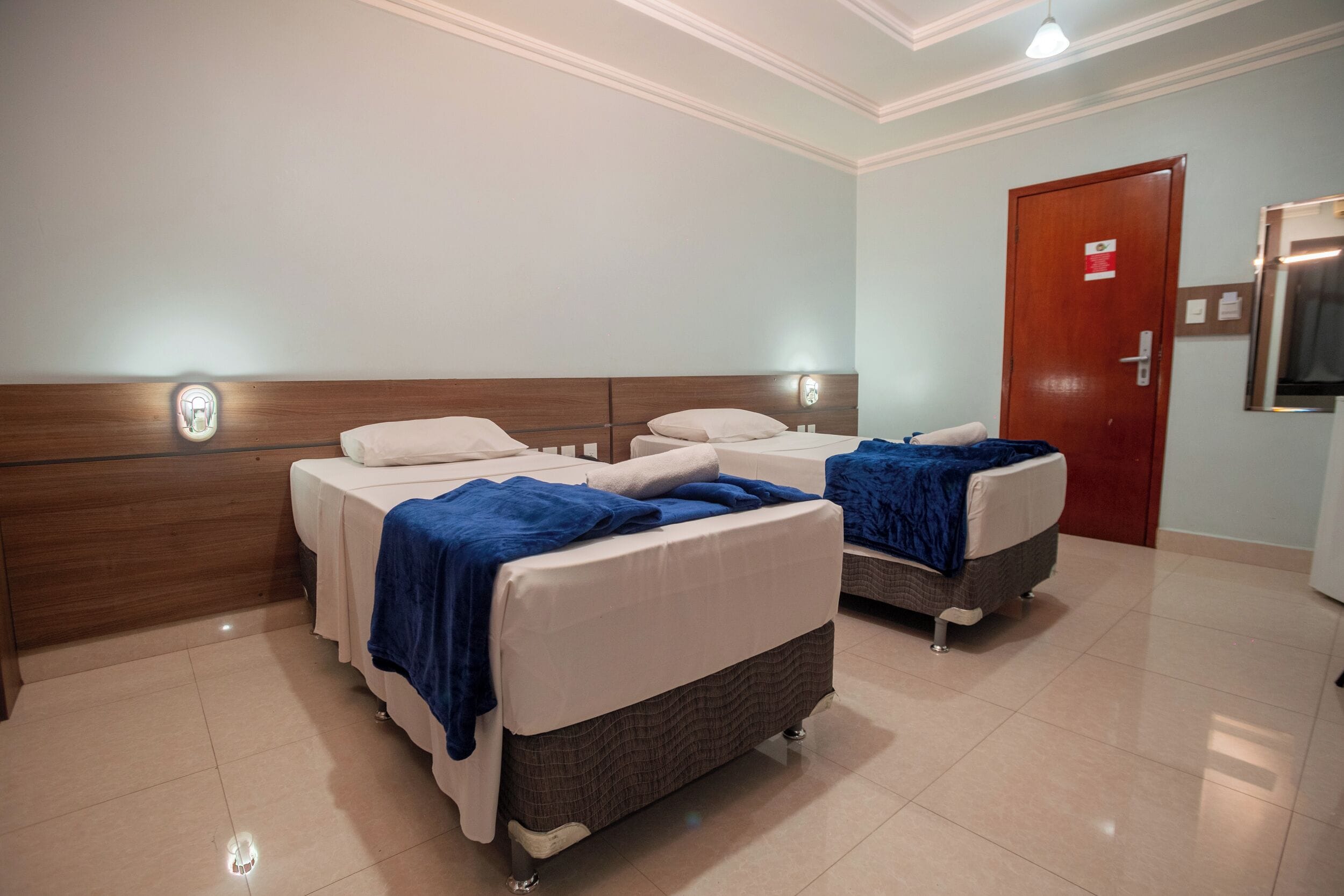 A hotel room with two beds, a wooden headboard, a door, and a wall-mounted light fixture.