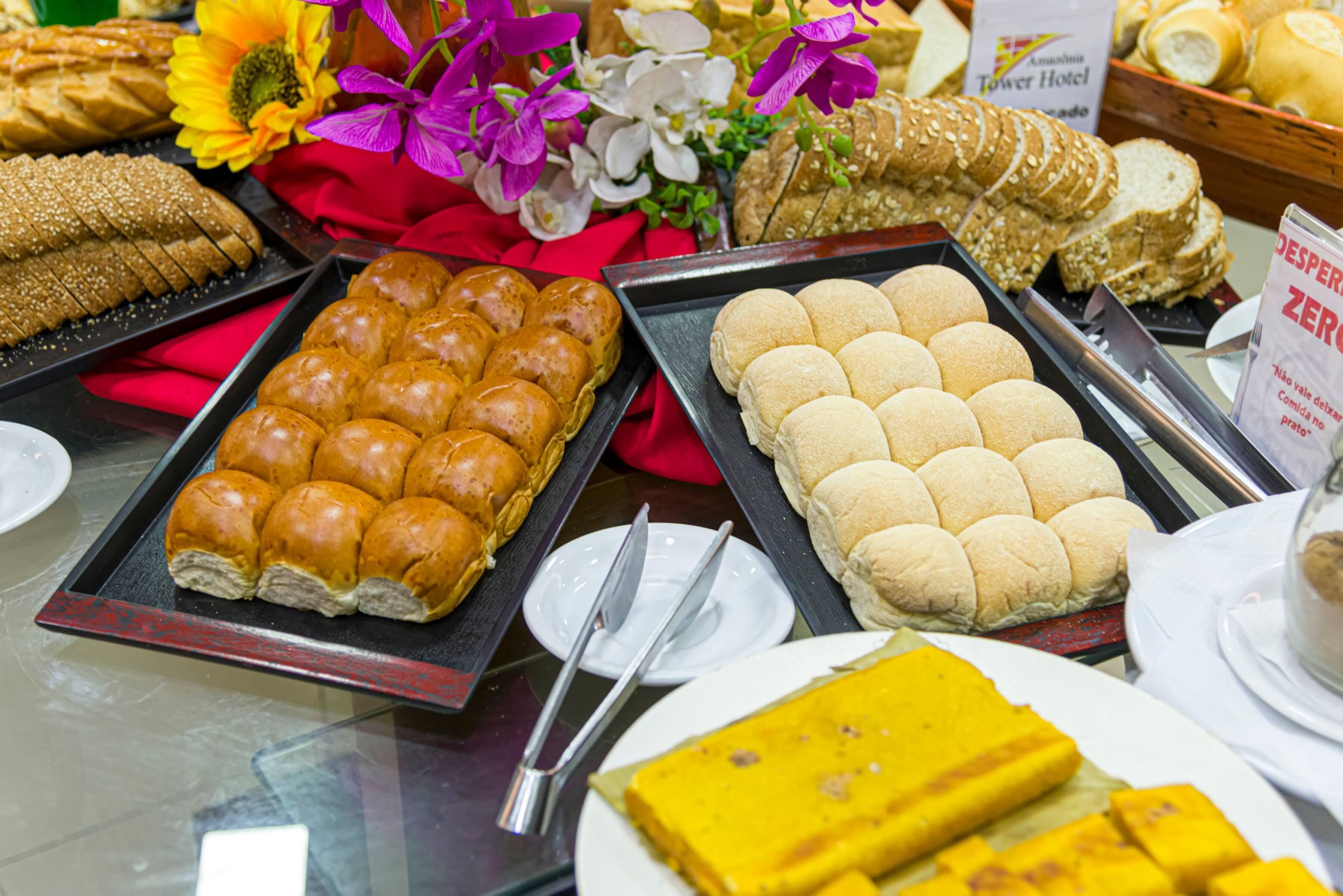 A buffet with various breads and pastries, including sesame seed rolls and dinner rolls, arranged on trays.