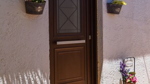 Una puerta marrón con un panel de vidrio, dos macetas de flores colgantes en la pared, un felpudo y una pequeña luminaria exterior.
