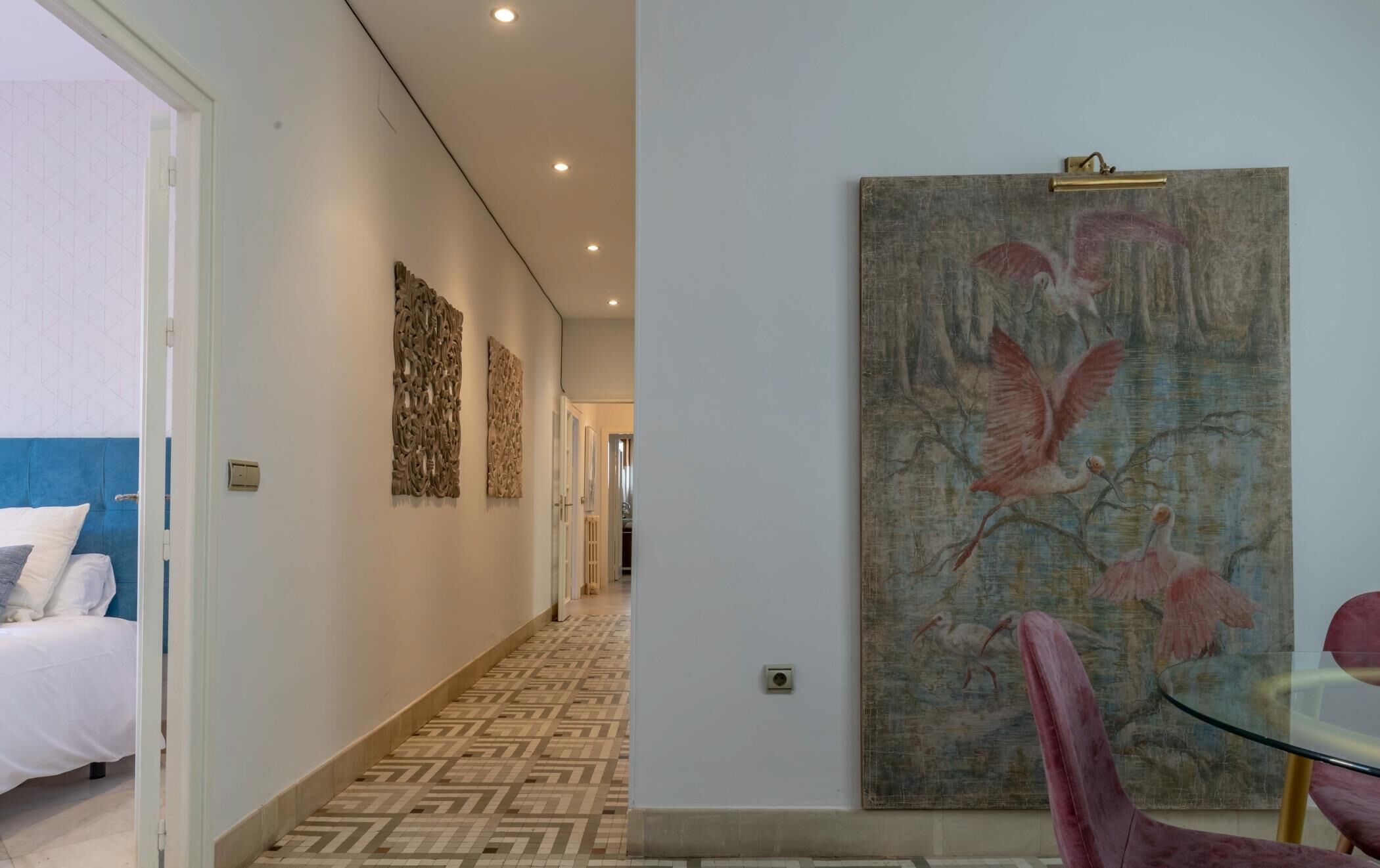 A hotel hallway with patterned flooring, a bed with white bedding, and a large painting of birds on the wall.