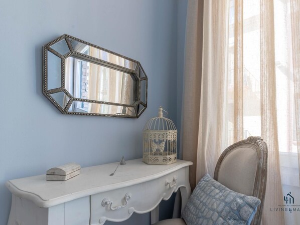 Una habitación con un tocador blanco, un espejo decorativo, una jaula de pájaros y una almohada azul estampada.