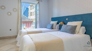 Ein Hotelzimmer mit zwei Betten, einem blauen Kopfteil und Blick auf Gebäude durch ein großes Fenster.