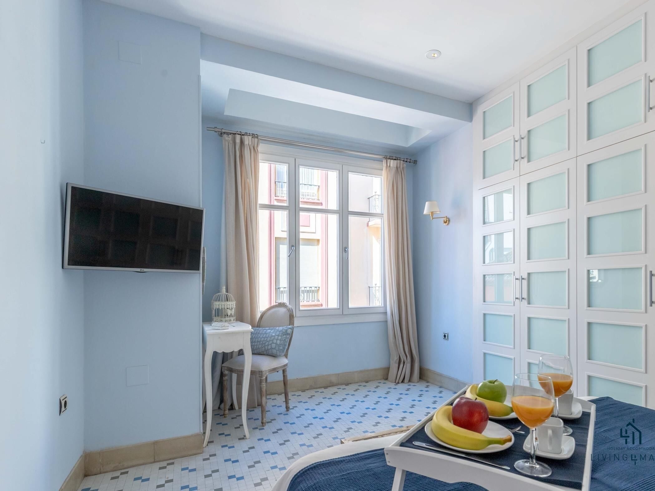 Une chambre avec un lit, une petite table sur laquelle se trouvent des fruits et des boissons, une chaise, un téléviseur et une fenêtre avec des rideaux.
