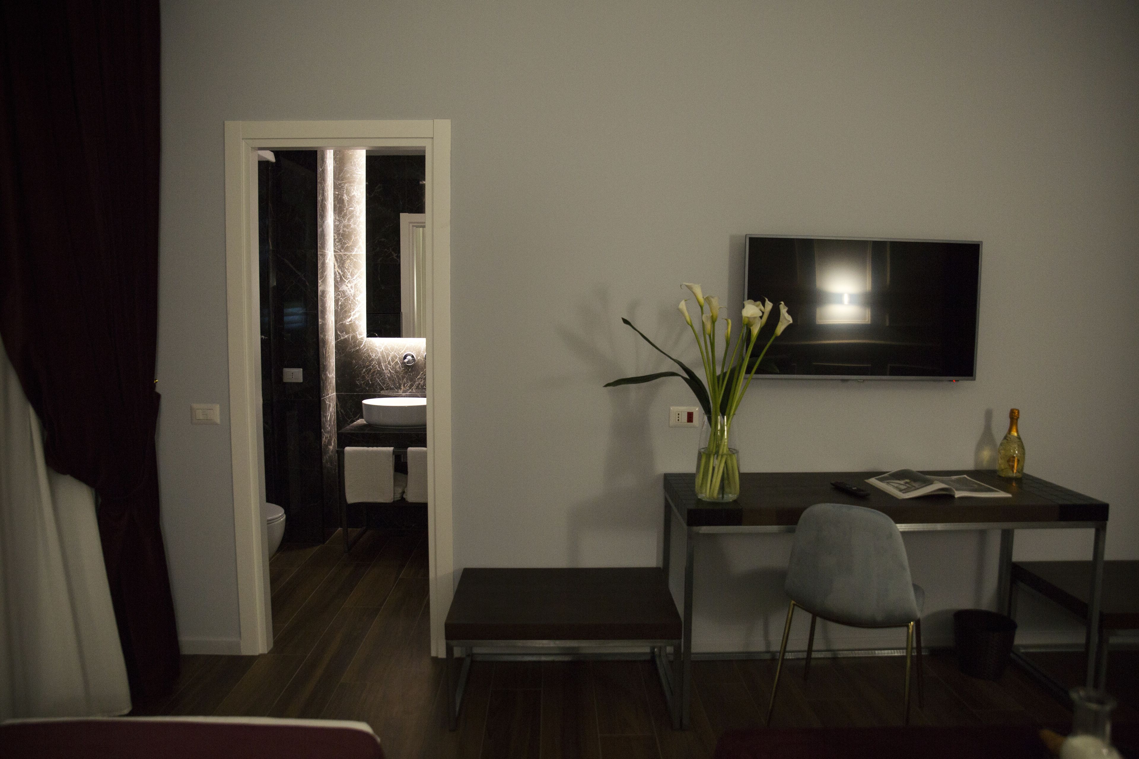 A modern living room with a dark wooden floor, a grey chair, a small table with a vase of flowers, a flat-screen TV mounted on the wall, and a doorway leading to a bathroom.