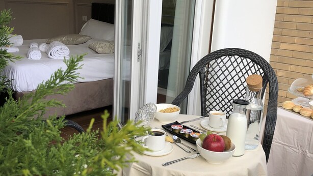 A balcony with a dining table set for breakfast, a bed with white linens, and a glass door leading to a room. - Unicum Roma Suites (Rome)