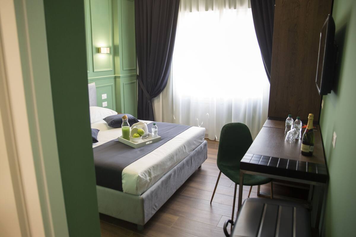 A hotel room with a bed, a dining table with a bottle and glasses, and a green chair.