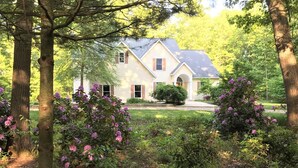 A two-story house with a gabled roof, surrounded by a well-maintained garden with flowering shrubs and a lawn. - Experience luxury at Riverwalk, a 4-bedroom retreat in serene Saugatuck, MI. (Saugatuck)