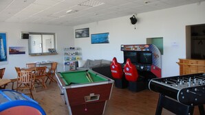 Una sala con mesa de billar, mesa de futbolín y mesa de pool. Hay sillas y una mesa con espejo.