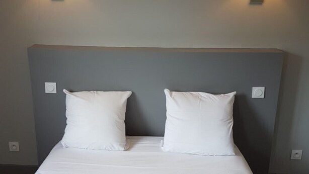 Una cama con ropa de cama blanca y dos almohadas, frente a una pared gris con dos interruptores de luz.