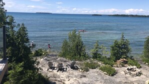 A lakeside view with kayaks, a rocky shore with Adirondack chairs, and distant trees.