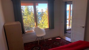 A room with a bed, a white chair, a window with curtains, and a view of trees and a body of water.