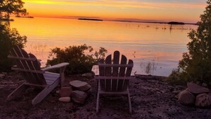 Two Adirondack chairs facing a sunset over a body of water.