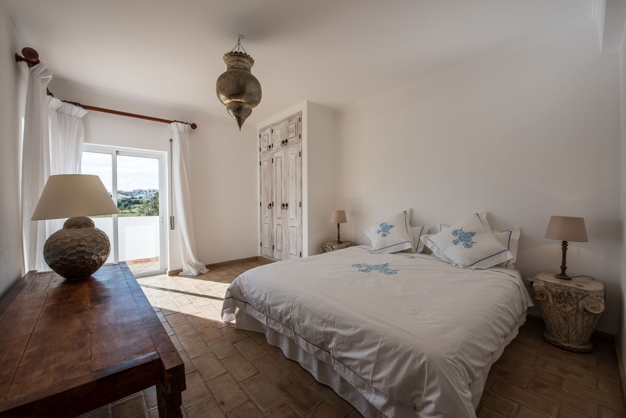 Un dormitorio con cama, una mesa de madera, lámpara, armario y una lámpara colgante.