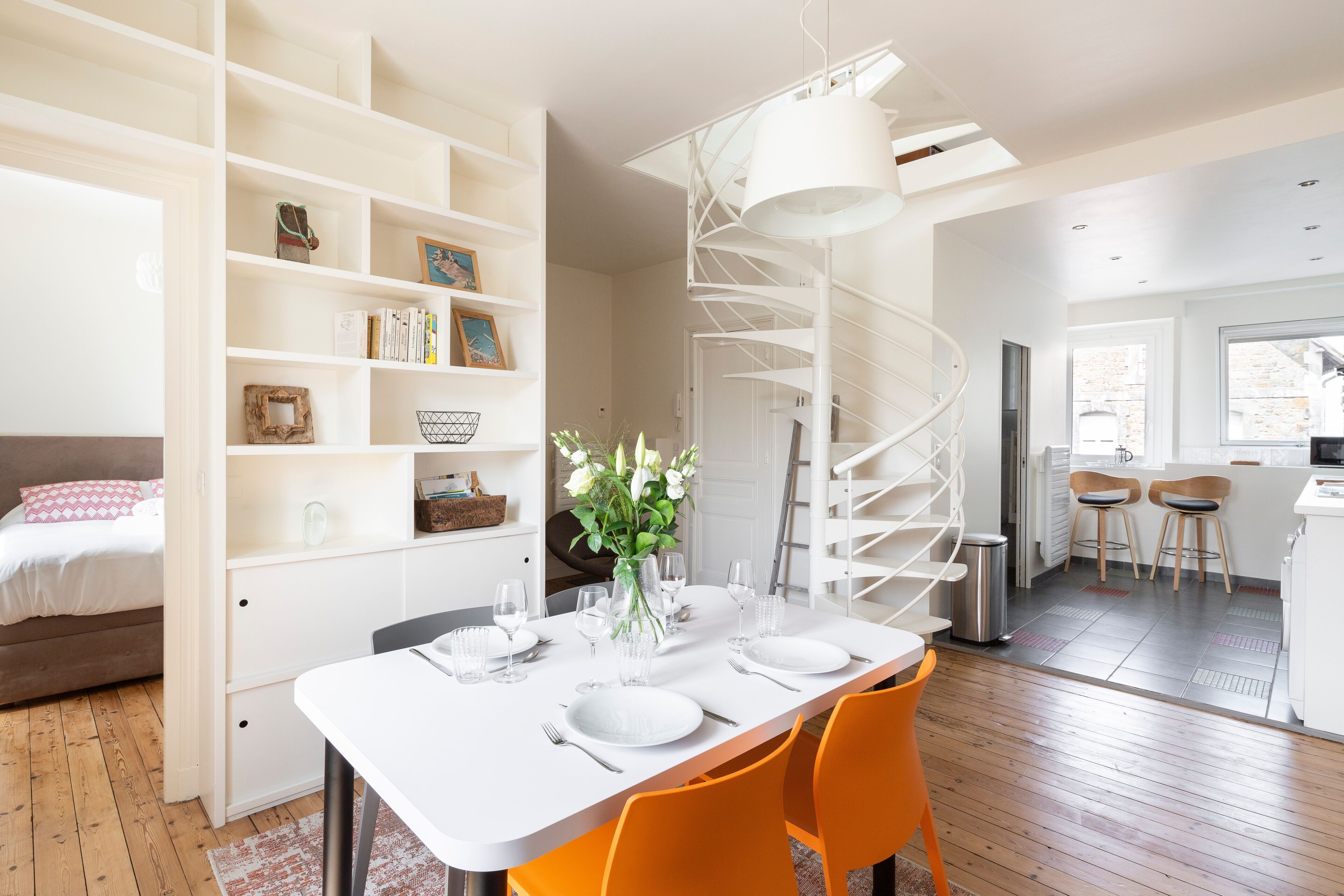 A modern interior with a spiral staircase, a dining area with a white table and orange chairs, and a kitchen with bar stools.