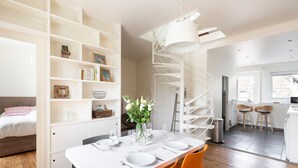 Un interior moderno con una escalera de caracol, un comedor con mesa blanca y sillas naranjas, y una cocina con taburetes de bar.
