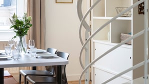 Un comedor con una mesa blanca, sillas negras, un jarrón con flores y una escalera de caracol con estantes blancos.