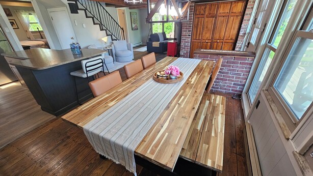 Une cuisine spacieuse avec une longue table en bois, un coin repas avec des chaises et une vue sur l’extérieur grâce à de grandes fenêtres.