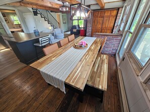 A spacious kitchen with a long wooden table, a dining area with chairs, and a view of the outdoors through large windows.