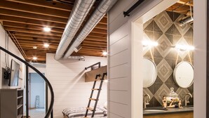 A modern loft bedroom with a loft bed, a wooden ladder, a black spiral staircase, and a bathroom with a geometric tile backsplash and round mirrors.