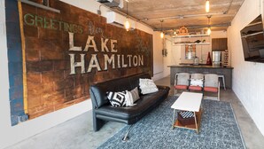 A modern living space with a black leather sofa, a white table, and a patterned rug. A brick wall with the text "Greetings from Lake Hamilton" is visible.