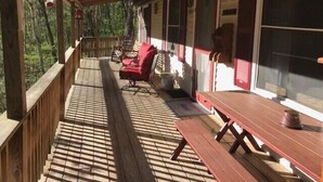 A covered porch with wooden railings, a red bench, a picnic table, and a view of the forest.