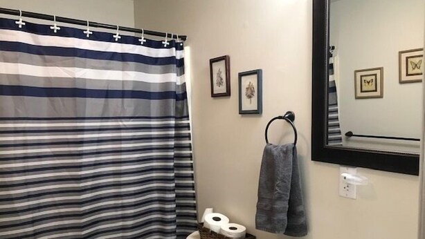 A bathroom with a striped shower curtain, a toilet with a basket of toilet paper, a towel rack with a towel, and a mirror with framed pictures on the wall.