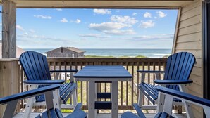 Um deck de madeira com uma mesa e quatro cadeiras Adirondack azuis, com vista para o oceano e um edifício ao fundo.