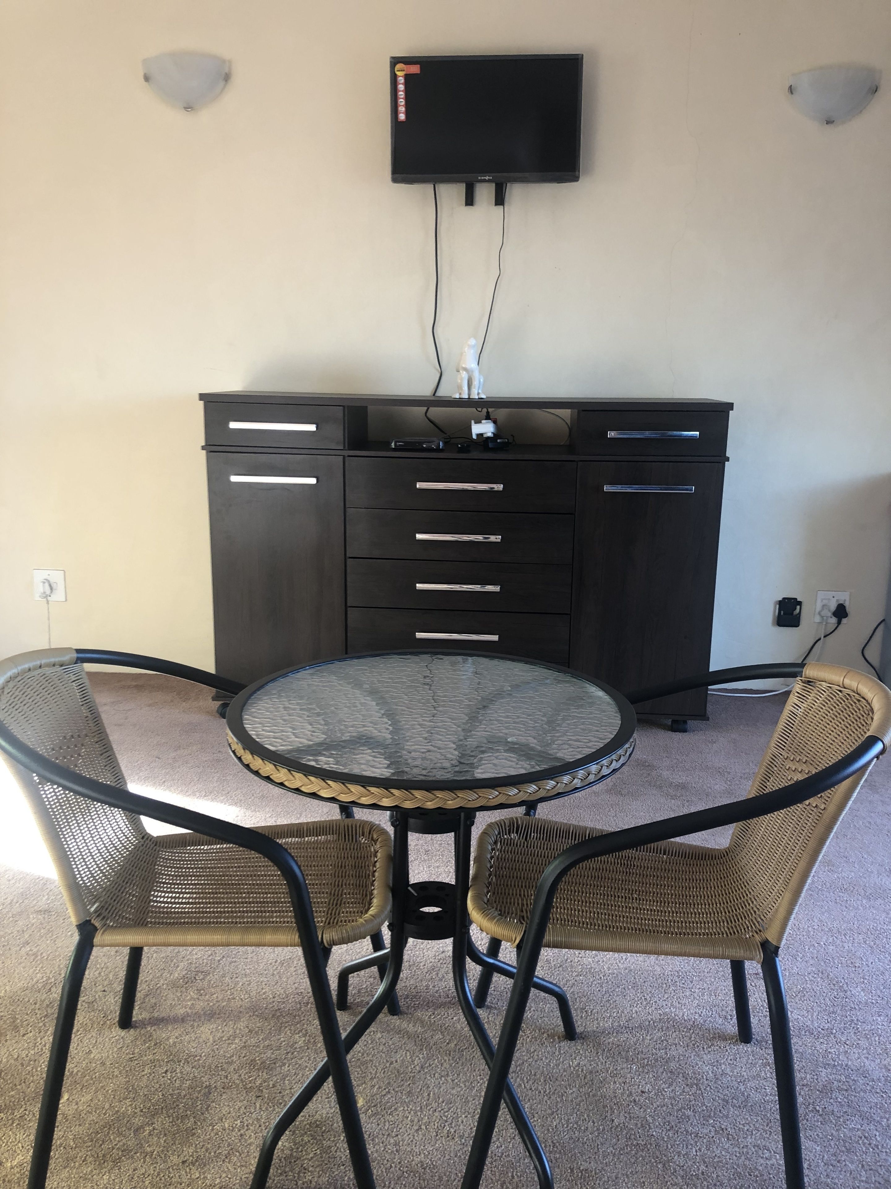 A room with a round glass table surrounded by wicker chairs, a dark wooden cabinet with drawers, and a mounted flat-screen TV.