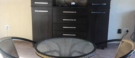 A room with a round glass table surrounded by wicker chairs, a dark wooden cabinet with drawers, and a mounted flat-screen TV.