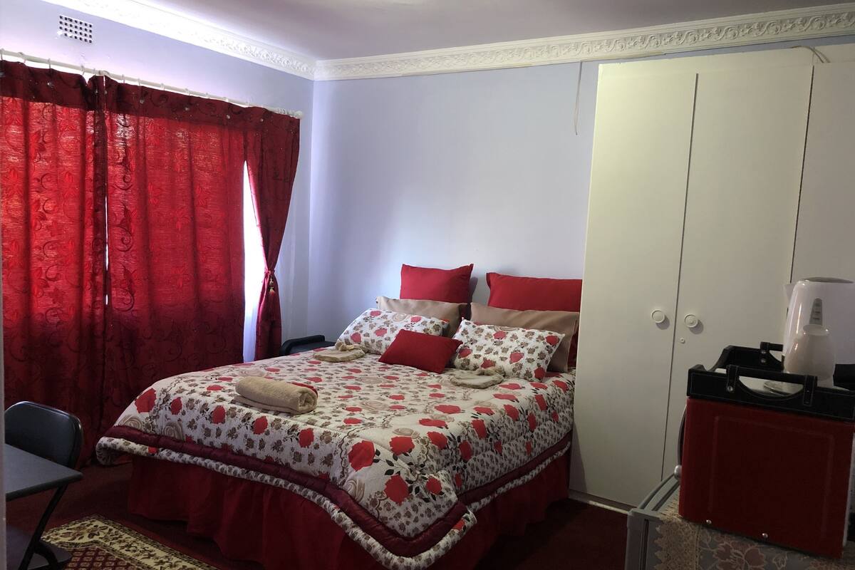 A bedroom with a bed, red curtains, a chair, a wardrobe, and a coffee maker.