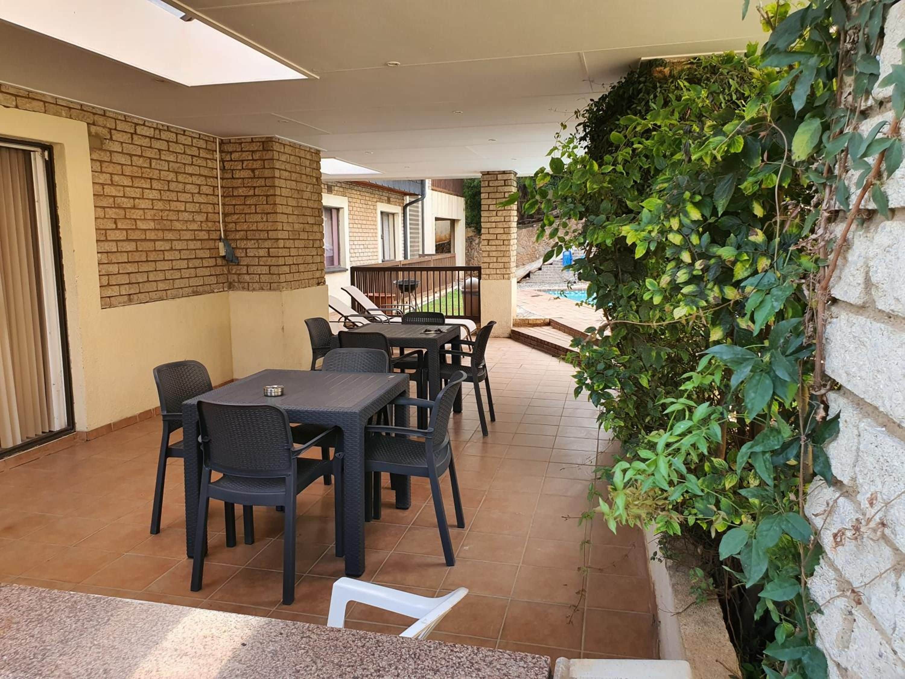 An outdoor patio area with a brick wall, a covered seating area with tables and chairs, and a view of a swimming pool in the background.