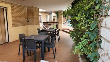 Un patio al aire libre con pared de ladrillo, una zona de estar cubierta con mesas y sillas, y vista a una piscina al fondo.