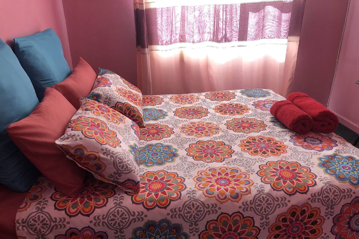 A bed with a colorful floral bedspread, two pillows, and a rolled towel.