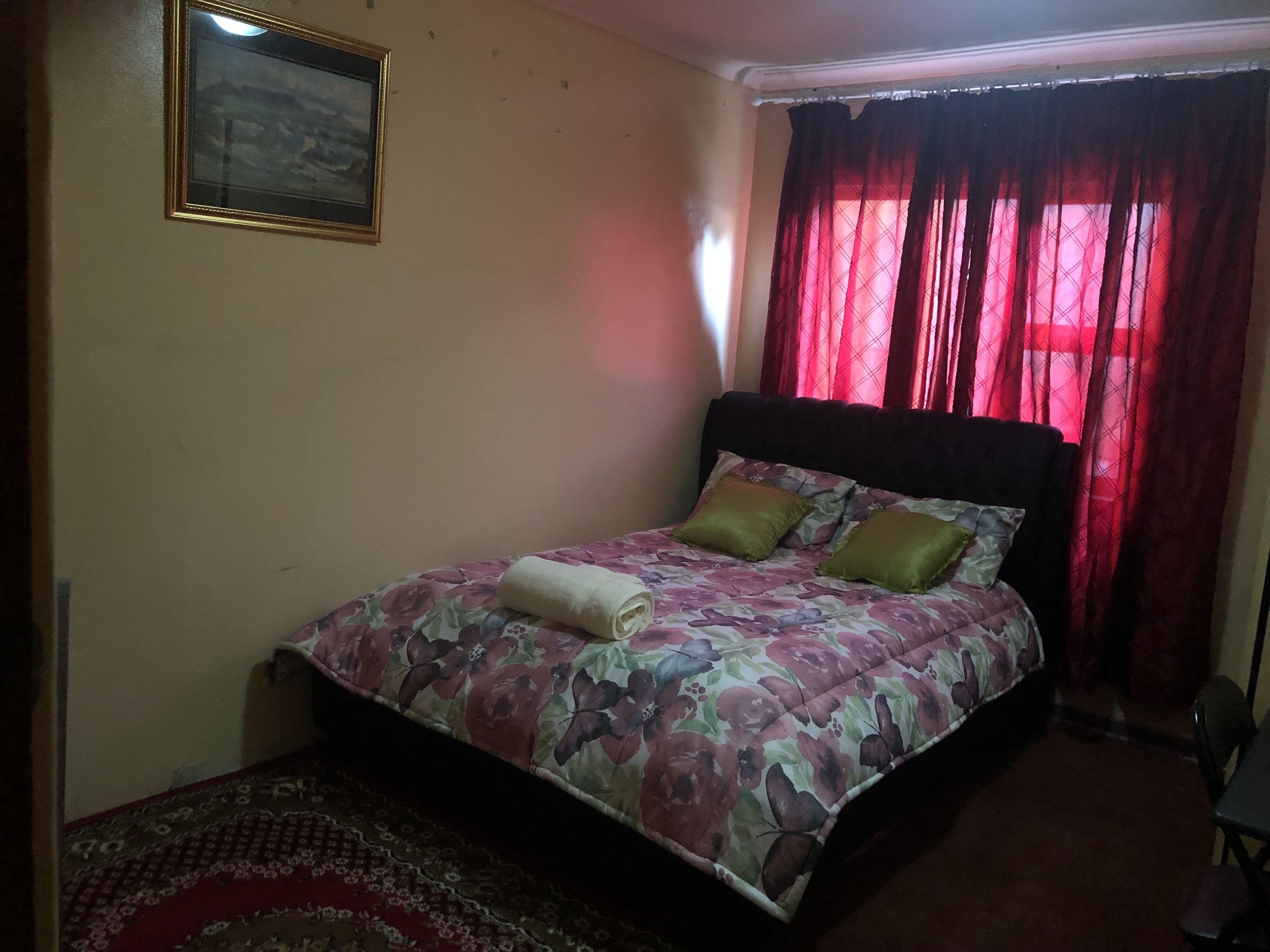 Un dormitorio con una cama, una ventana con cortinas rojas, un cuadro enmarcado en la pared y un tapete estampado.