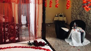 Una habitación de hotel con una cama, una silla, una mesa con una botella y una cortina con motivos florales.