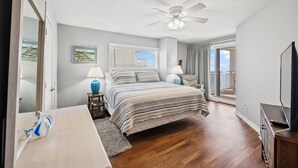 A bedroom with a bed, a desk, a chair, a lamp, a ceiling fan, and a view of the outdoors.