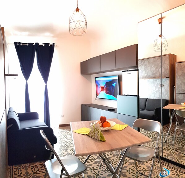 A modern living room with a sofa, a dining table with chairs, a television, and a kitchen area with cabinets. - Modular House Cefalù - Federica (Cefalù)