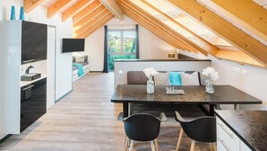 A modern loft apartment with a high ceiling, exposed wooden beams, a dining area with a black table and chairs, a kitchen with a built-in fridge, and a TV mounted on the wall. - 5* rooftop loft with roof terrace to enjoy /1-4 p/ garden use/parking/close to the city (Bensheim)