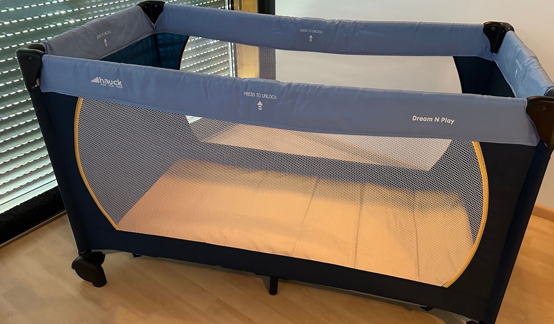 A baby playpen with a mesh floor and a padded top, labeled "Dream N Play" and "Press to Unlock".