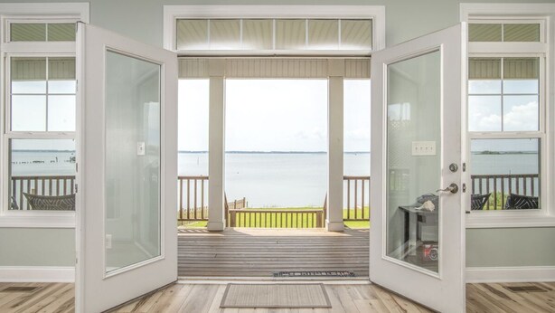 Une pièce avec vue sur un lac, accessible par de grandes portes vitrées, et donnant sur une terrasse en bois avec une petite table et une chaise.