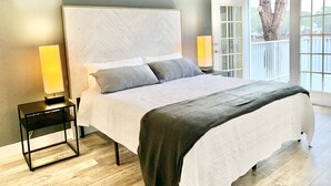 A modern bedroom with a large bed, two bedside lamps, a ceiling fan, and a view of the outdoors through a glass door.