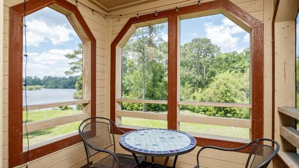 A covered outdoor seating area with a round table and chairs, large windows with wooden frames, and a view of a lake and trees. - Near beach indoor pool hot tub and outdoor pool (Myrtle Beach)