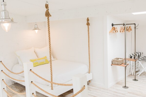 Una cama blanca con detalles en cuerda en una habitación luminosa, con una lámpara colgante y un perchero con ganchos.
