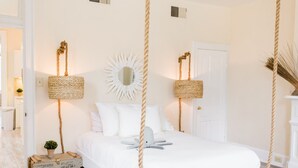 Ein Schlafzimmer mit einem weißen Bett, weißen Wänden, einer weißen Tür und einer weißen Decke. An der Decke hängen zwei Korbwäschekörbe, an der Wand ein weißer Spiegel und auf dem Boden ein kleiner Topfpflanze.