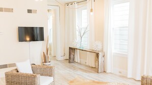 Ein modernes Wohnzimmer mit Fernseher, Korbmöbeln, einem Holztischchen und einem großen Fenster mit Vorhängen.