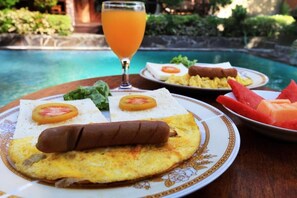 A breakfast setting with a plate of eggs, sausage, and tomato on a table by a pool. - Uma Dong Loka Village (Abiansemal)